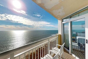 Een balkon met uitzicht op zee, een glazen deur die leidt naar een kamer met een bed en een nachtkastje, en een witte balustrade.