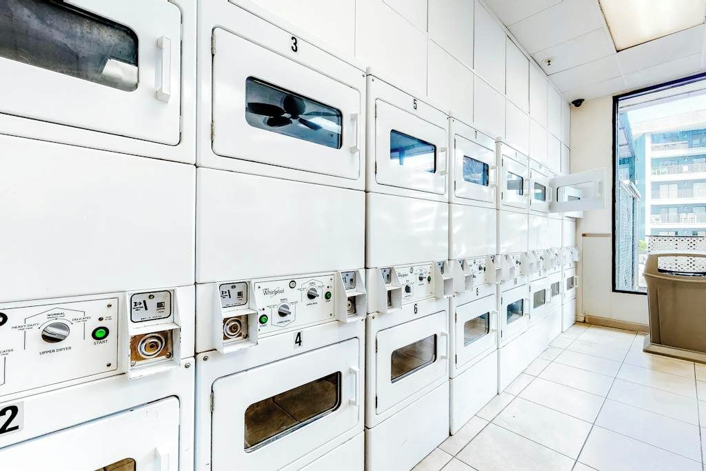 A row of white Whirlpool dryers with control panels, numbered from 2 to 6, in a well-lit laundry room with a large window.