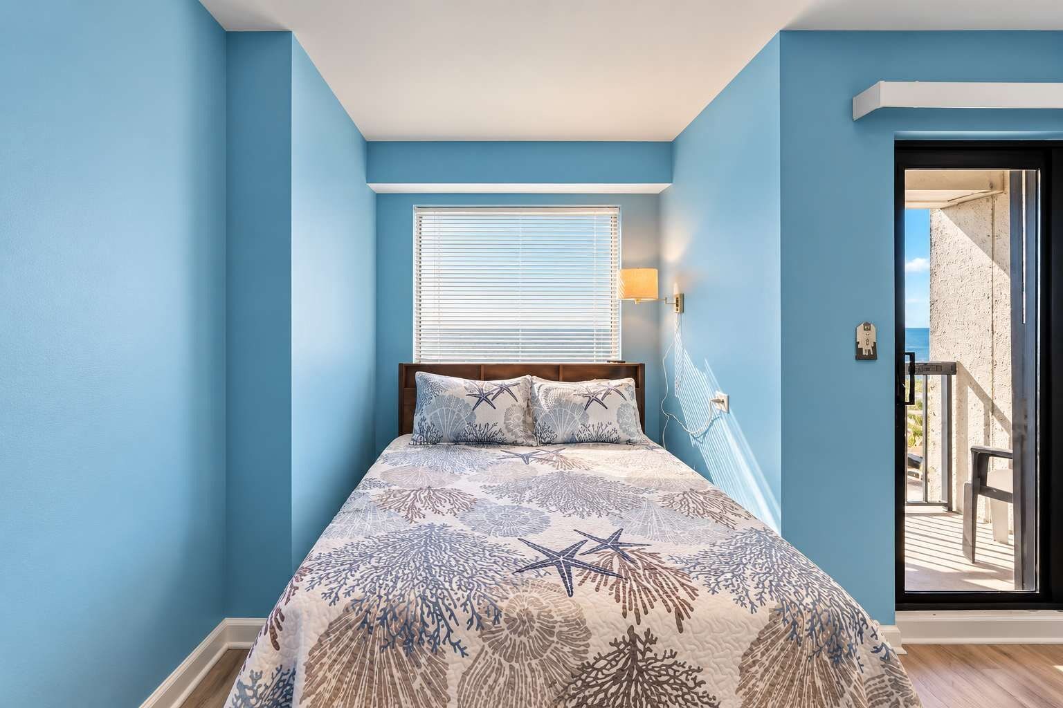 Une chambre à coucher avec un lit, une fenêtre avec des stores, une porte donnant sur un balcon et un luminaire fixé au mur.