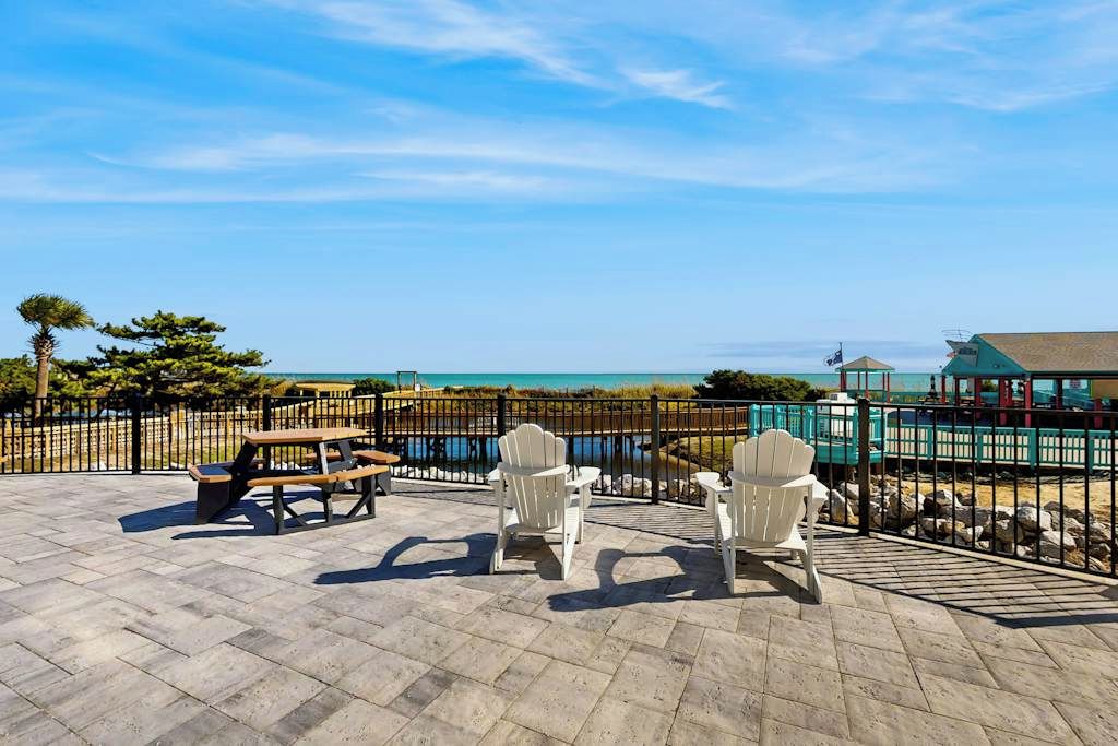 An outdoor patio with picnic tables and Adirondack chairs, overlooking a beach and a body of water.