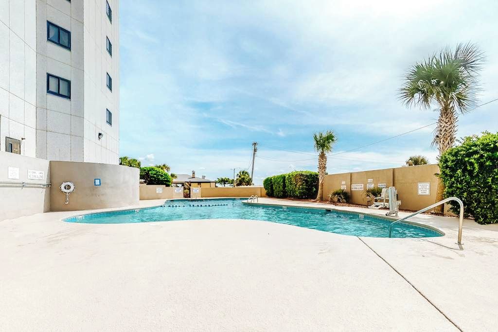 A swimming pool with a pool ladder, surrounded by a low wall and palm trees.