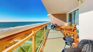 Ein Balkon mit Blick auf Strand und Meer, ein Tisch mit Stühlen und ein Fenster mit Vorhängen.
