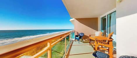A balcony with a view of the beach and ocean, a table with chairs, and a window with curtains.