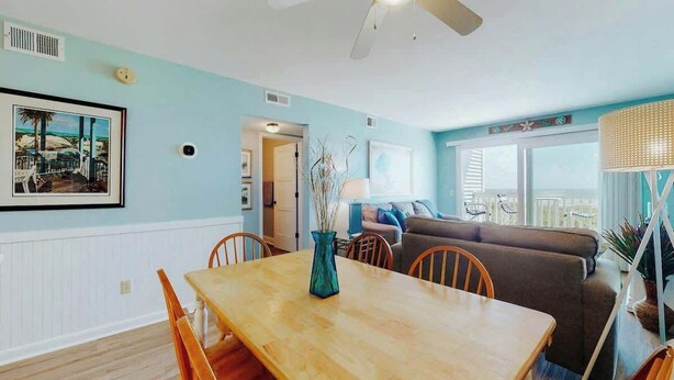 A dining area with a wooden table and chairs, a sofa, and a lamp in a room with light blue walls. - Ocean Retreat Unit N1 (North Myrtle Beach)