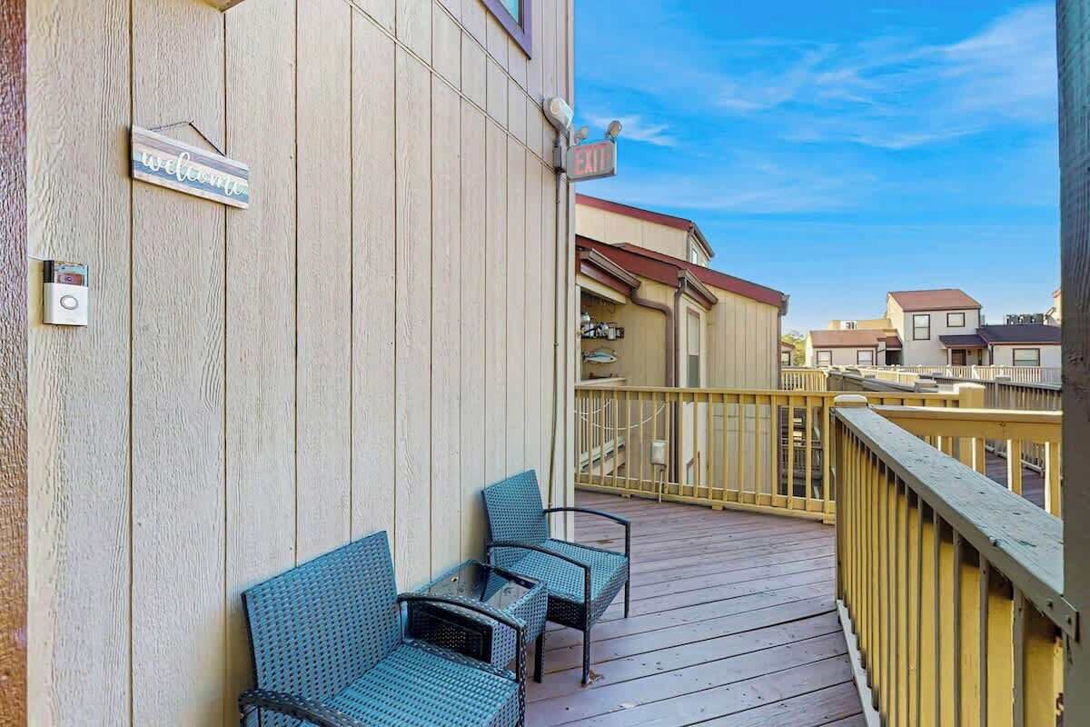 Una terraza de madera con dos sillas de mimbre y una pequeña mesa, un letrero de bienvenida y un letrero de salida en un edificio.