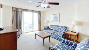 Ein Hotelzimmer mit Deckenventilator, einer Couch, einem Couchtisch, einem Schreibtisch und einem Fenster mit Vorhängen.
