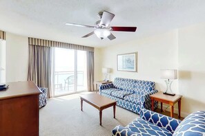 Ein Hotelzimmer mit Deckenventilator, einer Couch, einem Couchtisch, einem Schreibtisch und einem Fenster mit Vorhängen.