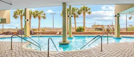 An indoor swimming pool with clear views of palm trees and a wooden deck in the background.