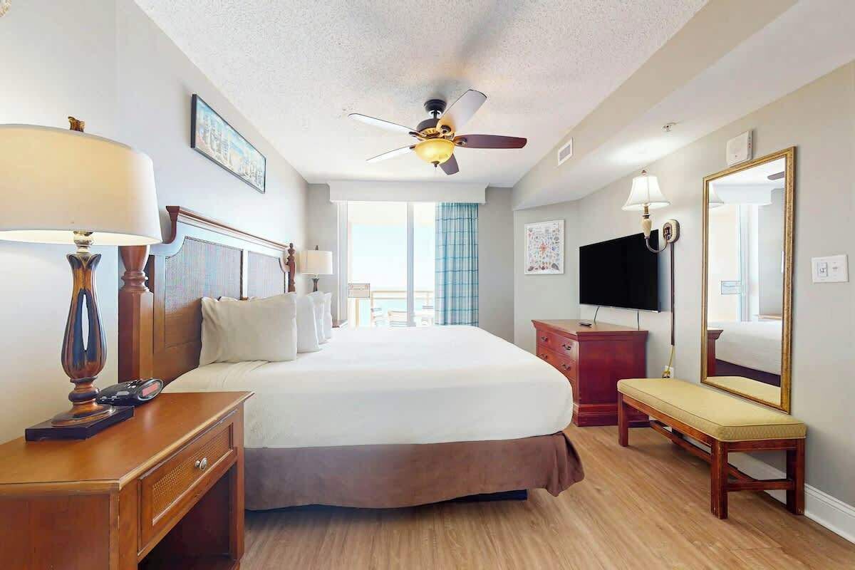 A hotel room with a bed, bedside table, lamp, dresser, bench, ceiling fan, and a TV.