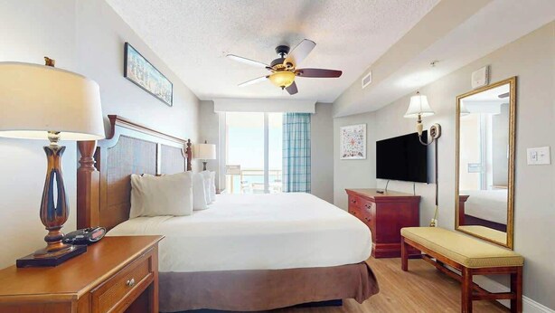 A hotel room with a bed, bedside table, lamp, dresser, bench, ceiling fan, and a TV. - Bahama Sands Unit 0707 (North Myrtle Beach)