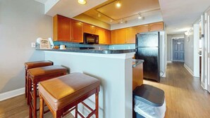 A modern kitchen with wooden cabinets, a black refrigerator, and a bar area with stools. - Bahama Sands Unit 0707 (North Myrtle Beach)
