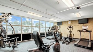 Et fitnesscenter med forskellig træningsudstyr, store vinduer og et fjernsyn på væggen.
