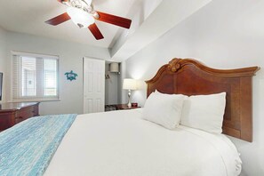 Une chambre d’hôtel avec un grand lit, un ventilateur de plafond, une fenêtre avec des stores, une porte et une petite table avec une lampe.