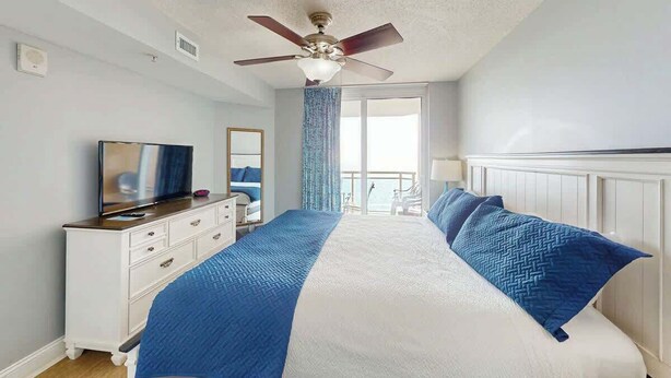 Une chambre à coucher comprenant un lit, une commode, un téléviseur, un ventilateur de plafond et une fenêtre avec des rideaux.