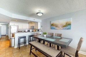 A modern kitchen with a central island, a dining area with a long table and benches, and a painting on the wall. - Bahama Sands Unit 0806 (North Myrtle Beach)