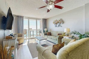 A living room with a ceiling fan, a flat-screen TV, a sofa, a coffee table, and a view of the outdoors. - Bahama Sands Unit 0807 (North Myrtle Beach)