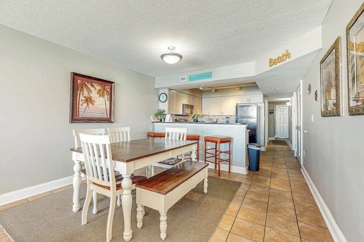 A dining area with a wooden table and chairs, a kitchen in the background, and a hallway leading to other rooms.