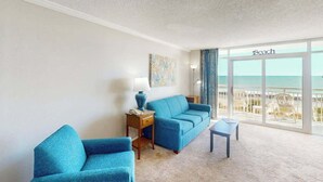 Een kamer met een groot raam met uitzicht op een strand, een blauwe bank, een tafeltje en een lamp.