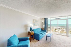 Ein Zimmer mit großem Fenster mit Blick auf einen Strand, ein blaues Sofa, ein kleiner Tisch und eine Lampe.