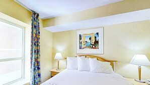 Ein Hotelzimmer mit einem Bett, zwei Nachttischlampen, einem Fenster mit Vorhängen und einem gerahmten Bild an der Wand.