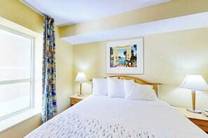 Ein Hotelzimmer mit einem Bett, zwei Nachttischlampen, einem Fenster mit Vorhängen und einem gerahmten Bild an der Wand.