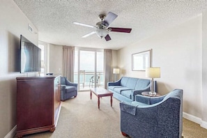 Ein Hotelzimmer mit Deckenventilator, einem großen Fenster mit Vorhängen, einem Sofa, einem Sessel, einem kleinen Tisch und einem Fernseher auf einem Schrank.