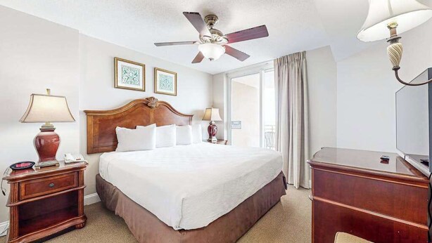 A hotel room with a bed, bedside tables, a ceiling fan, a television, and a window with curtains. - Bahama Sands Unit 0905 (North Myrtle Beach)
