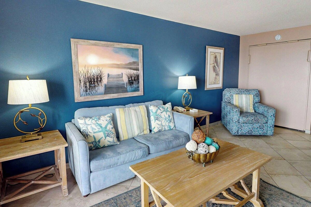 Un salon avec un mur bleu, un canapé, un fauteuil à motifs, une table basse en bois et deux petites tables d’appoint avec des lampes.