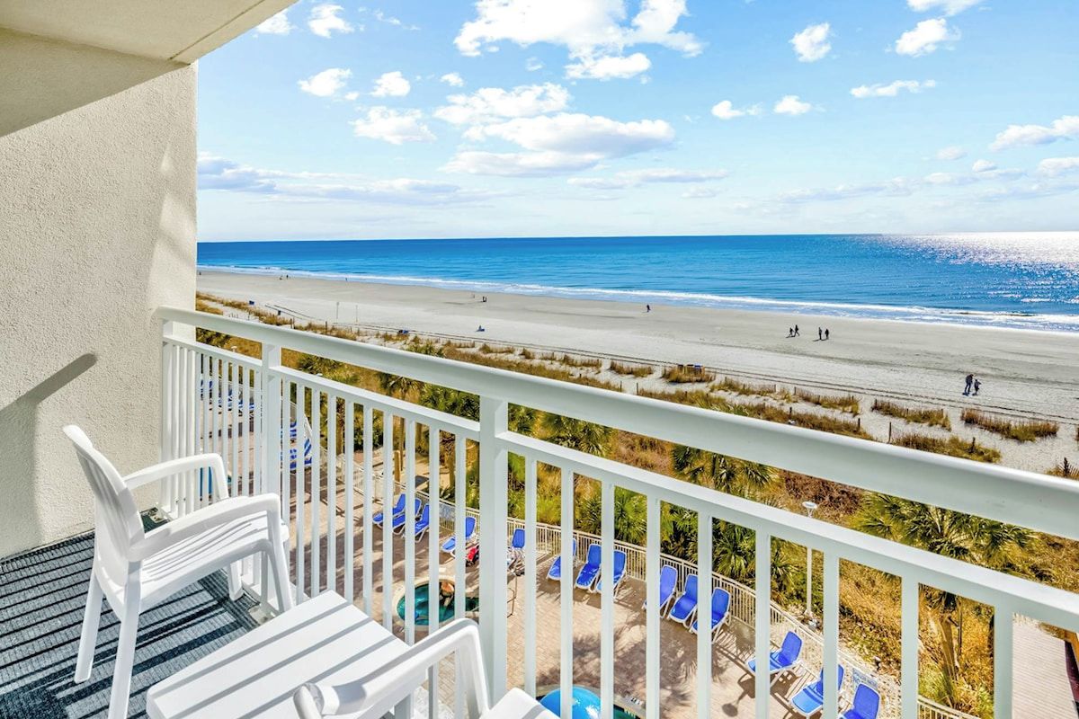 A balcony with a view of a beach, white chairs, and a pool area.