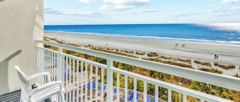 A balcony with a view of a beach, white chairs, and a pool area.