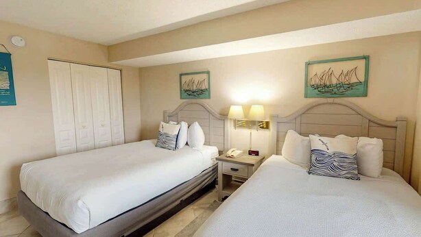 Quarto de hotel com duas camas, um guarda-roupa, mesas de cabeceira, luminárias e obras de arte nas paredes.