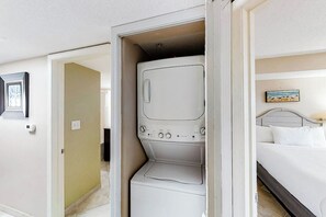 Quarto de hotel com uma cama, uma máquina de lavar e uma porta que dá para outro cômodo.
