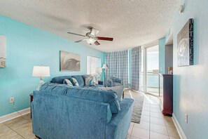 Ein Zimmer mit blauem Sofa, Deckenventilator, Vorhängen am Fenster und Blick ins Freie.
