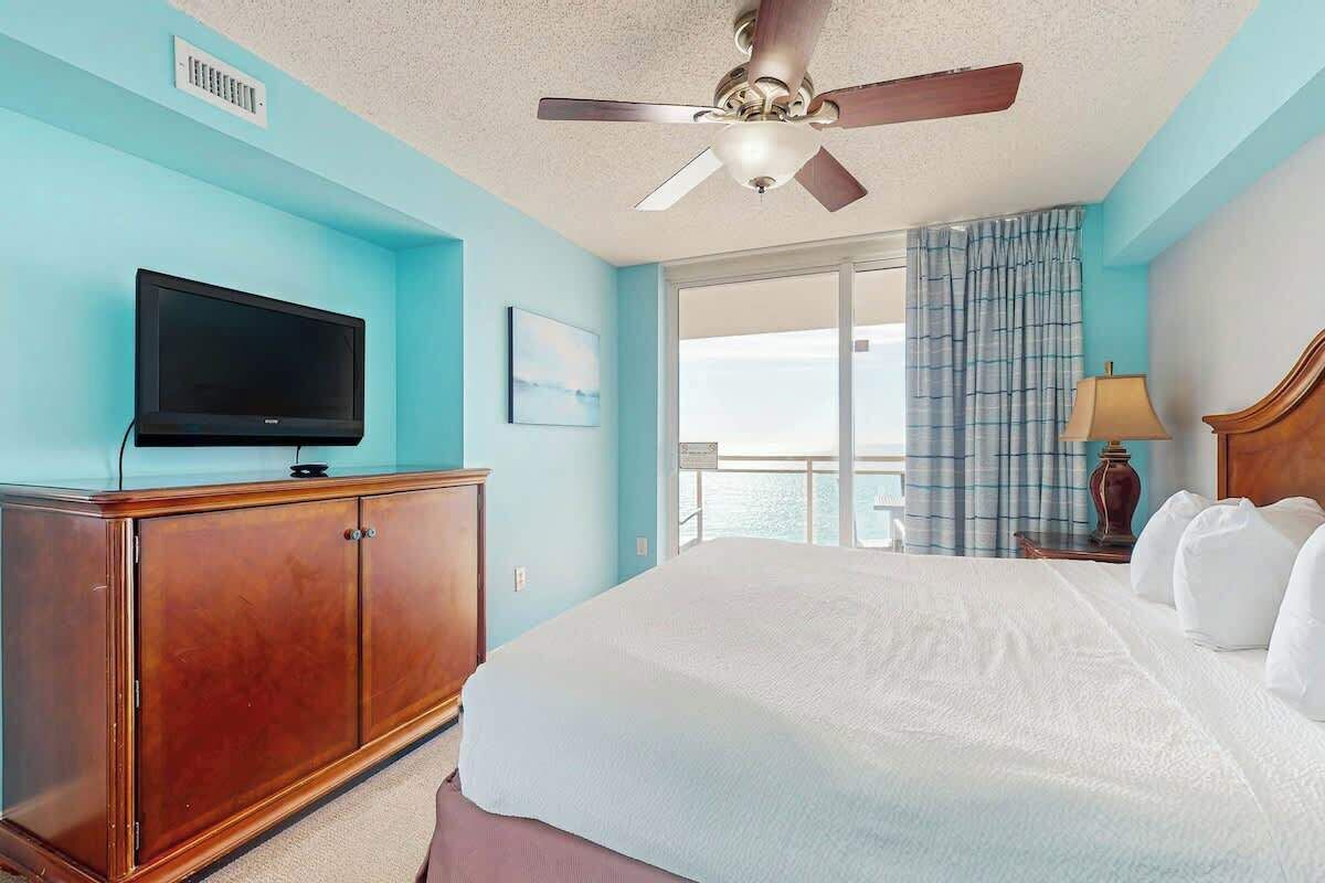A hotel room with a bed, a television on a wooden cabinet, a ceiling fan, and a view of the sea through a window.