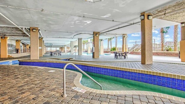 En indendørs swimmingpool med flisekant, røde mursten i gulvet og et overdækket område med borde og stole.