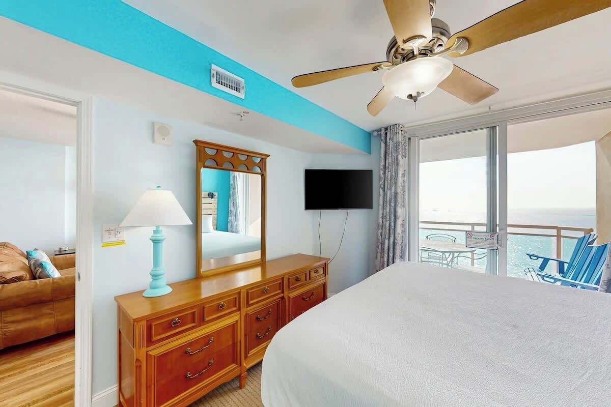 Habitación de hotel con cama, cómoda, ventilador de techo, televisión y balcón con vistas.