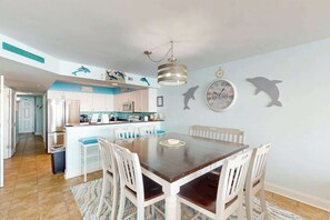 A dining area with a wooden table and white chairs, a kitchen in the background, and dolphin-themed wall decorations. - Bahama Sands 0906 - Dolphin's Delight (North Myrtle Beach)