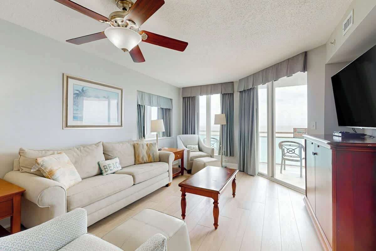 A hotel room with a sofa, armchairs, a coffee table, a TV, and a balcony with curtains.