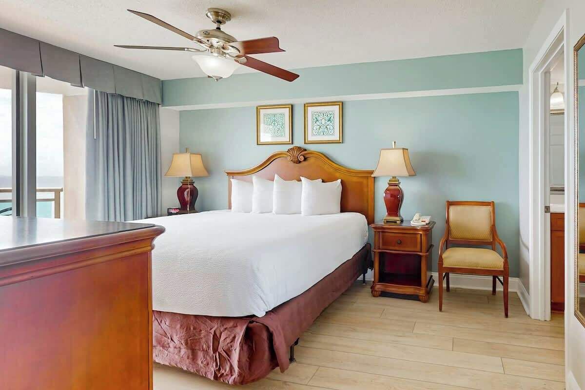 Ein Hotelzimmer mit einem großen Bett, zwei Nachttischen, einem Sessel, einem Deckenventilator und einem Fenster mit Vorhängen.