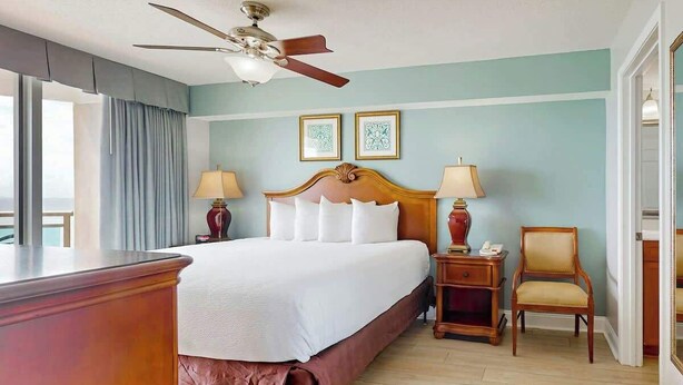A hotel room with a large bed, two bedside tables, a chair, a ceiling fan, and a window with curtains. - Bahama Sands Unit 1002 (North Myrtle Beach)