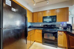 A kitchen with wooden cabinets, a black refrigerator, a microwave, and a stove. - Bahama Sands Unit 1002 (North Myrtle Beach)