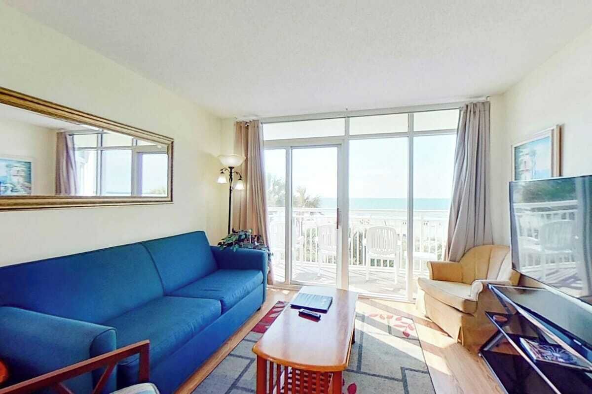 A room with a blue sofa, a wooden coffee table, a TV, and a balcony with a view.