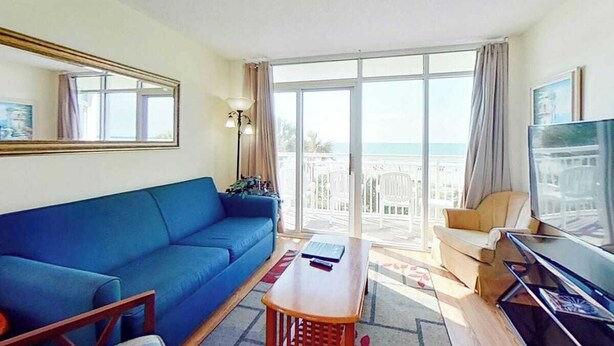 A room with a blue sofa, a wooden coffee table, a TV, and a balcony with a view. - Bay Watch Resort 0202 (North Myrtle Beach)