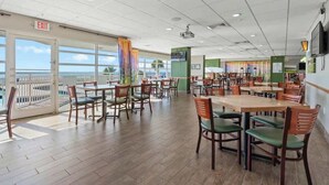 A spacious dining area with multiple tables and chairs, large windows offering a view, and an exit sign on the wall. - Bay Watch Resort 0202 (North Myrtle Beach)