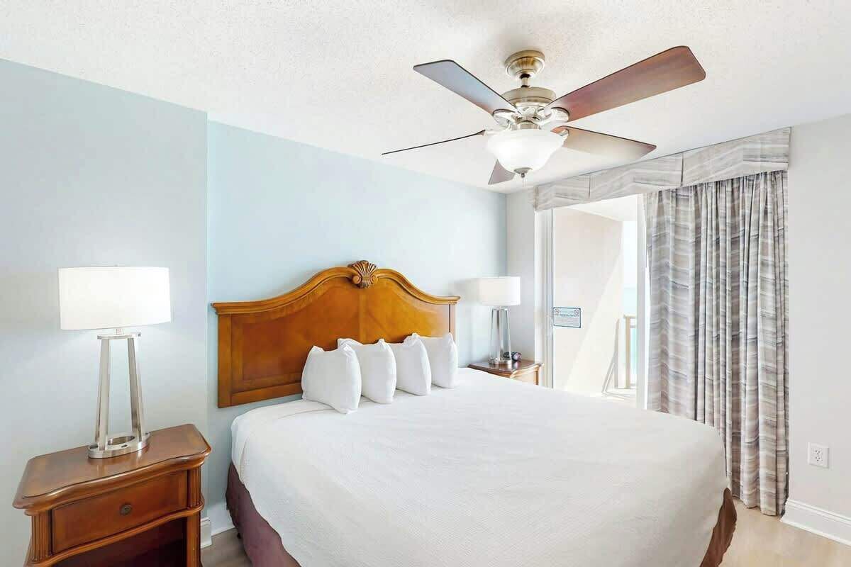 Une chambre d’hôtel avec un lit, une table de chevet, une lampe, un ventilateur de plafond et une fenêtre avec des rideaux.