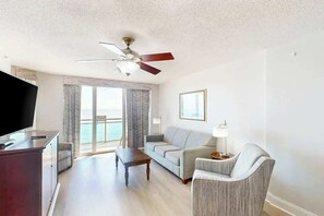 A hotel room with a television, a sofa, a chair, a coffee table, a ceiling fan, and a view of the ocean through large windows. - Bahama Sands Unit 1003 (North Myrtle Beach)