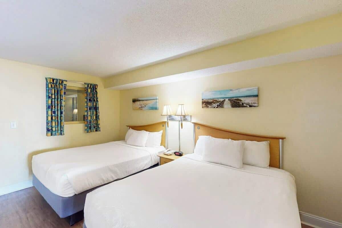 A hotel room with two single beds, a bedside table with a lamp, a mirror, and two framed pictures on the wall.