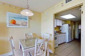 A dining area with a table and chairs, a kitchen in the background, and a beach-themed wall decoration. - Bay Watch Resort 0206 (North Myrtle Beach)