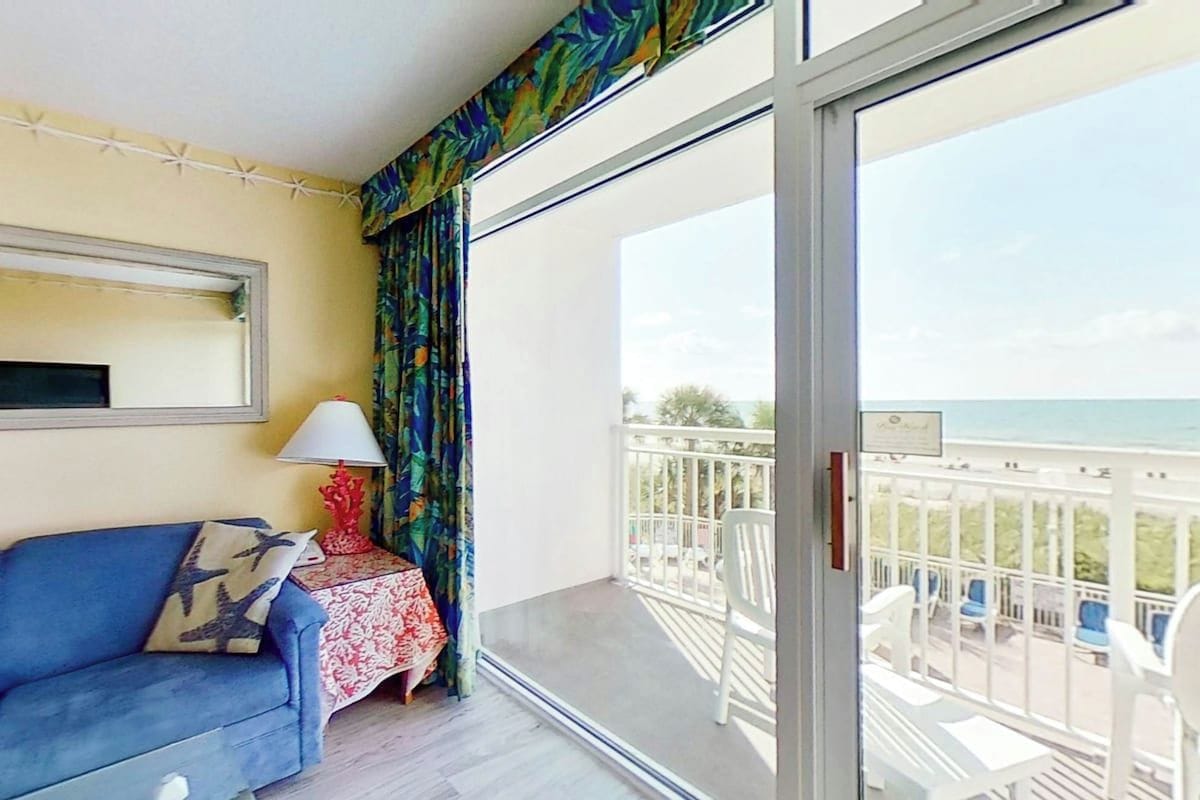 A balcony with a view of the sea, a blue sofa, a table with a red lamp, and a patterned curtain.