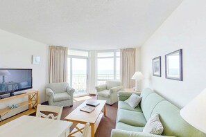 A hotel room with a television, two armchairs, a sofa, a dining table, and a balcony with curtains. - Bay Watch Resort 0412 Oceanfront (North Myrtle Beach)